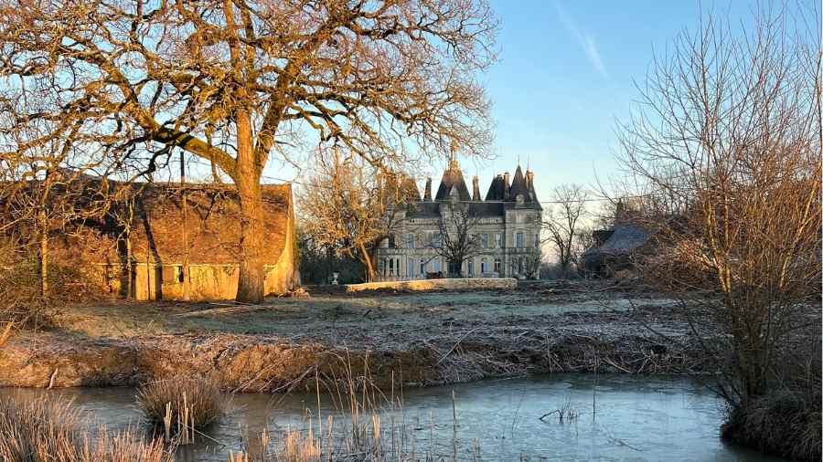 Wide view of Château de la Boutinière from across the water, with the historic château buildings framed by trees and winter landscape.