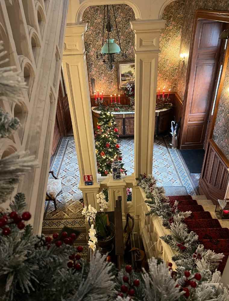 View down the château staircase decorated with Christmas garlands, lanterns and a lit tree visible in the hall below.