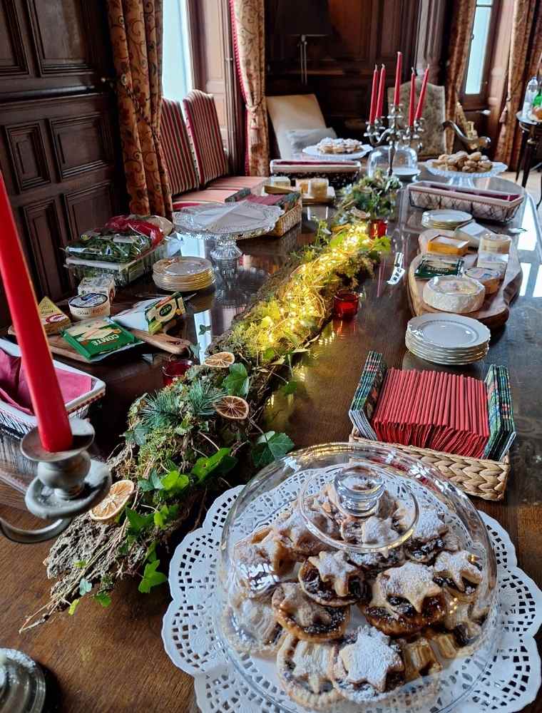 A long dining table laid with mince pies, plates and napkins, decorated with greenery, candles and lanterns for a Christmas gathering.