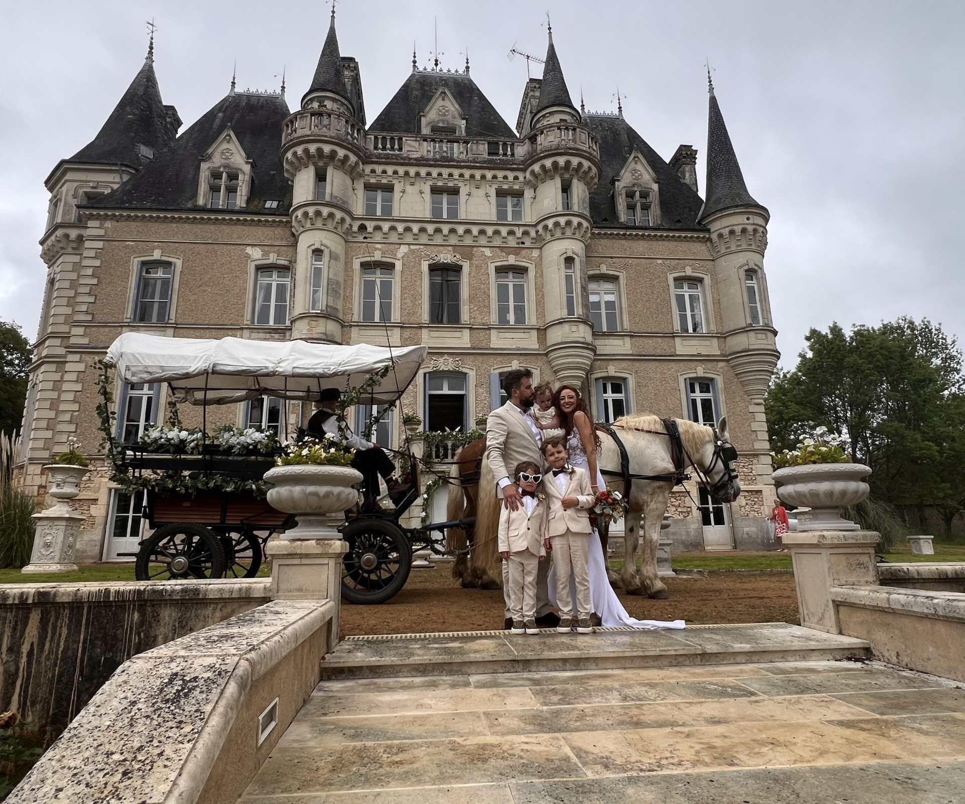 Wedding couple with two children standing in front of Chateau de la Boutinière, with a horse-drawn carriage and château façade behind them, showcasing the grounds as a micro wedding venue in France.