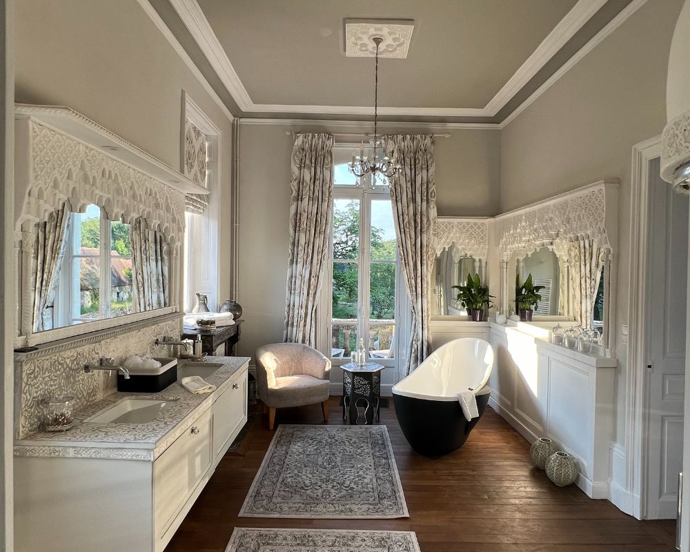 Elegant bridal suite bathroom with carved white detailing, twin sinks, chandeliers, a freestanding black-and-white bath, wooden flooring, and tall French windows overlooking the gardens at Château de la Boutinière.