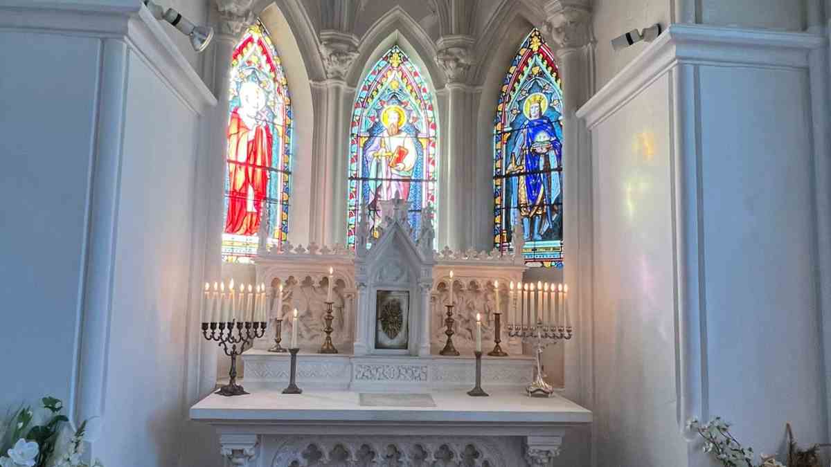 White carved altar inside the chapel at Château de la Boutinière, lit by candles and framed by three tall stained glass windows.
