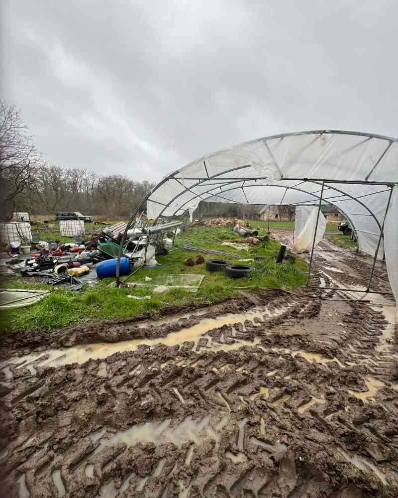 The greenhouse stands twisted and partly displaced after the storm, surrounded by deep mud, tyre tracks and scattered gardening equipment.