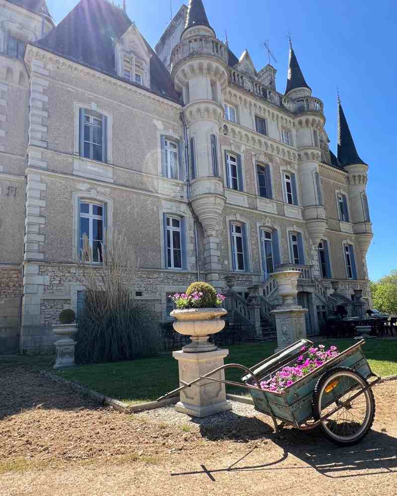 Chateau events gardening day showing a wide view of the château façade with flower-filled urns and a cart planted with pink flowers.